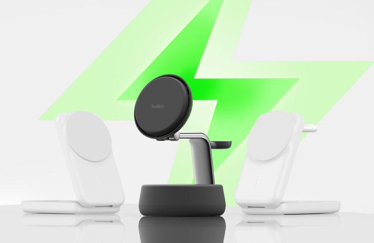 Three Belkin wireless charging stands displayed on a glossy surface, with a large green lightning bolt graphic in the background symbolizing fast charging. The center stand is black with a sleek, elevated design, while the two on either side are white with a more compact, upright style.