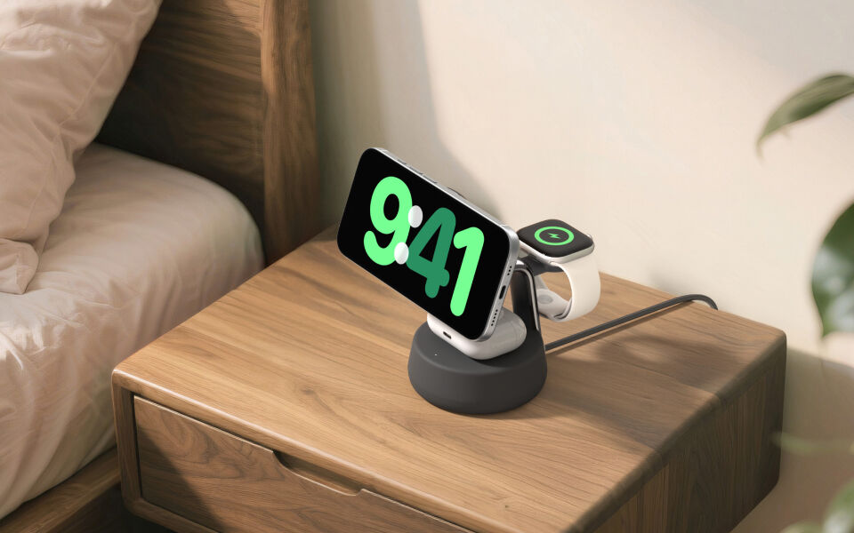 Black 3-in-1 wireless charging stand on a wooden bedside table, charging a smartphone in landscape mode, a smartwatch, and wireless earbuds.