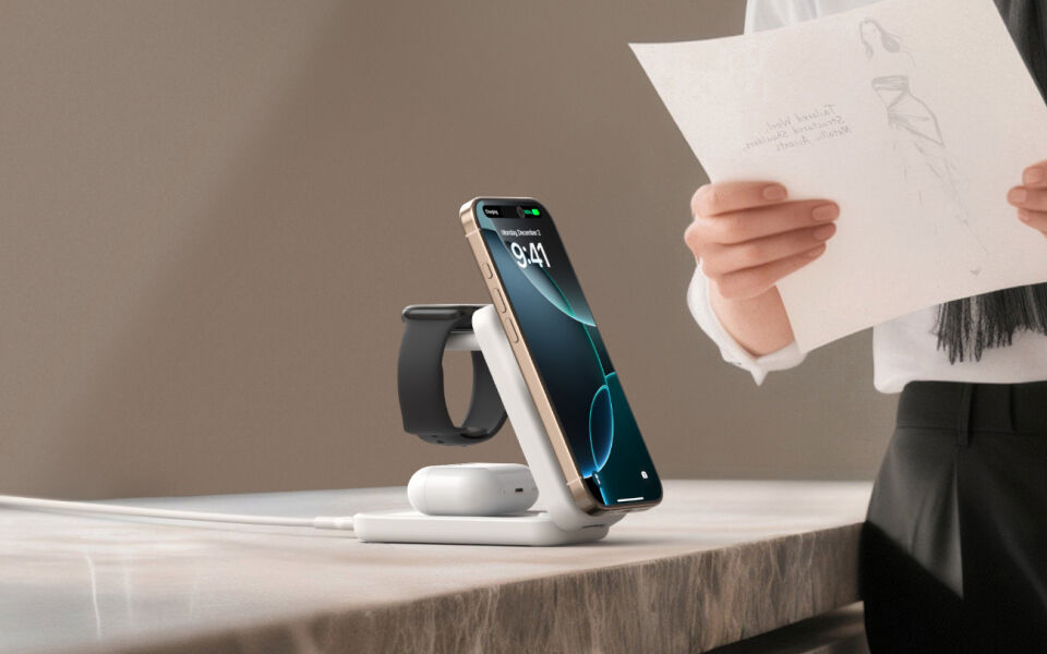 White Belkin 3-in-1 wireless charging stand on a marble surface, charging a smartphone, smartwatch, and wireless earbuds, with a person in the background holding a sketch.