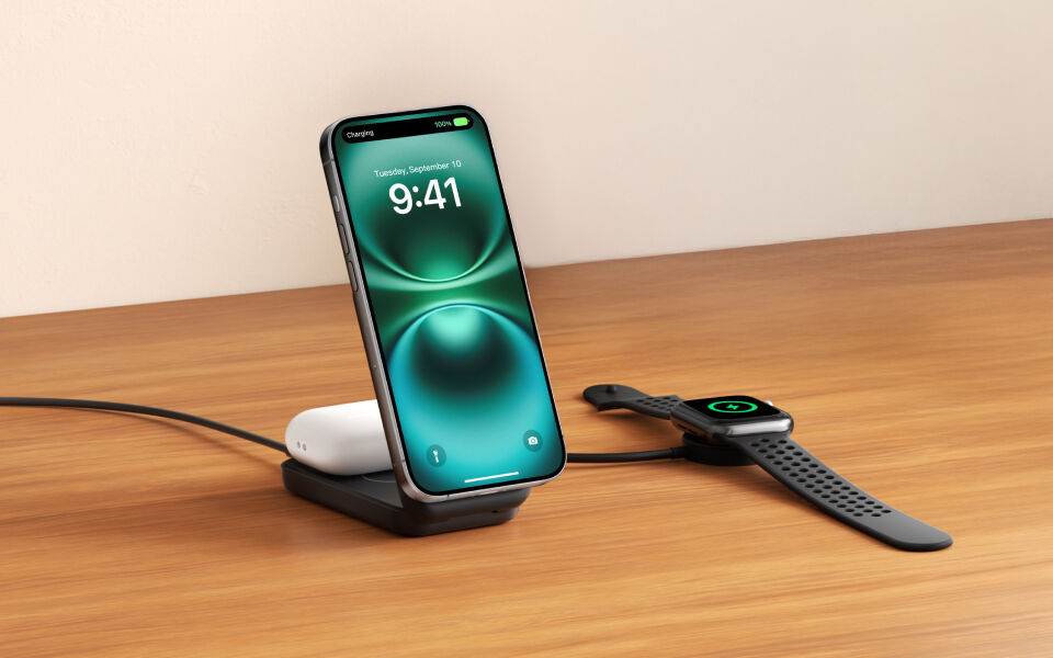 Black Belkin dual wireless charging stand on a wooden surface, charging a smartphone and wireless earbuds, with a smartwatch charging separately on a connected puck.