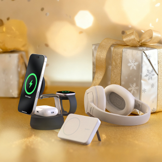 Holiday gift scene with Belkin tech products, including a wireless charging stand holding a phone and smartwatch, over-ear headphones, and a charging pad, placed in front of wrapped presents with gold ribbons.