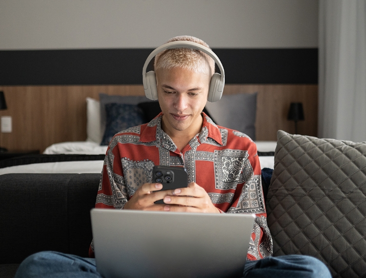 The image shows a young man with short, light-colored hair wearing over-ear headphones. He is sitting in a modern, comfortable room, possibly a bedroom or a lounge, with a bed and cushions visible in the background. He is smiling slightly while looking at his smartphone, which he holds in both hands, with a laptop placed in front of him on his lap. He is wearing a patterned red shirt.