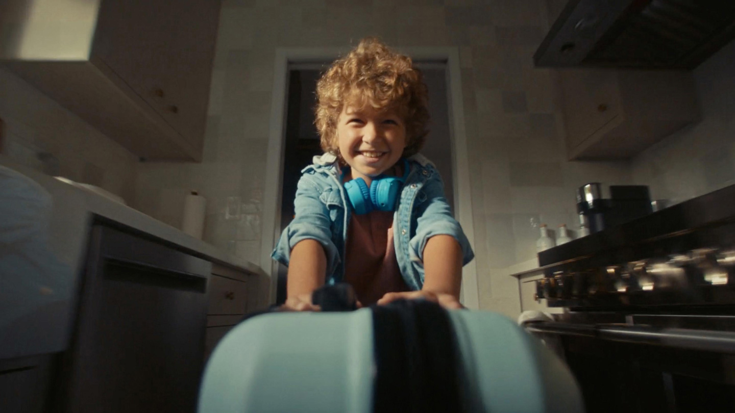 身穿牛仔外套、頸項上戴著一副亮藍色耳機的捲髮男孩微笑著，在廚房朝相機方向推著一個藍色行李箱。