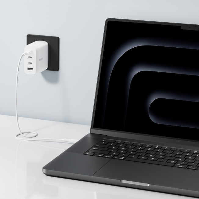 A laptop connected to a white Belkin wall charger plugged into a black power outlet on a light-colored wall. The charger has multiple ports and a white cable connecting it to the laptop, which displays a dark abstract design on its screen. The setup rests on a clean white desk.