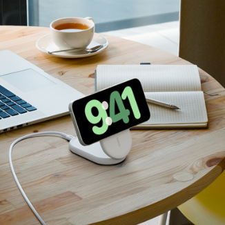 This image shows a wireless charging stand on a round wooden table, holding a smartphone in an upright position. The phone's screen displays the time as 9:41. The setup includes a connected charging cable, a laptop, an open notebook with a pen, and a cup of tea or coffee, creating a productive and cozy workspace environment. The scene is well-lit, with natural light from a nearby window, emphasizing a clean and organized aesthetic.