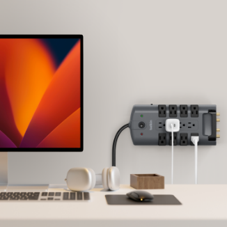 A Belkin surge protector mounted on a wall above a minimalist desk setup, powering multiple devices including a desktop computer, with headphones and accessories neatly arranged nearby.