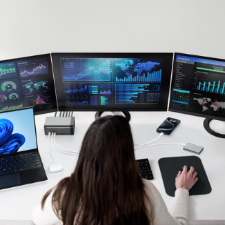 Person working at a desk with a laptop and three large monitors displaying data dashboards, connected through a docking station with multiple devices including a smartphone.